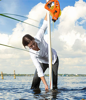 A person stands in shallow water, using equipment to measure or study the water. The sky is partly cloudy, and there is water and vegetation visible in the background.