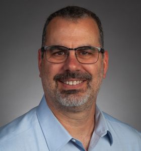 A man with short dark hair, glasses, and a gray beard wearing a light blue collared shirt smiles against a gray background.