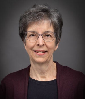 A woman with short gray hair, glasses, and a maroon cardigan over a black top, smiling slightly in front of a gray background.