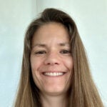 A woman with long brown hair smiles at the camera in front of a plain light-colored wall.