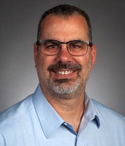 A middle-aged man with short hair, glasses, and a beard is smiling at the camera. He is wearing a light blue collared shirt against a plain gray background.