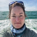 A woman wearing sunglasses on her head and a neck gaiter takes a selfie on a boat with the ocean and waves in the background.