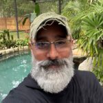 A man with a gray beard, glasses, and a green cap takes a selfie in front of a backyard pool surrounded by plants and trees.