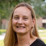 A woman with straight blonde hair, wearing a black top, smiles at the camera outdoors with greenery and buildings in the background.