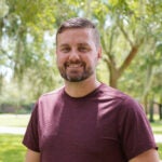 A man with short hair and a beard, wearing a maroon t-shirt, stands outdoors in a park with trees and sunlight in the background.