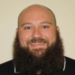 A smiling bald man with a full, dark beard, wearing a black shirt with "ucf" logo, against a light beige background.