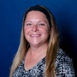 Woman with long blonde hair smiles, wearing a black and white patterned top and a black headband, against a blue background.
