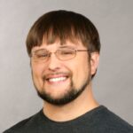 A person with short hair, glasses, a beard, and a mustache, wearing a dark shirt, smiling against a plain background.