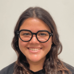 A person with long brown hair, wearing round black glasses and a black shirt, smiling in front of a plain light-colored background.