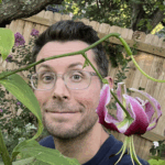 A man with glasses stands in a garden, smiling next to a large purple and white lily flower, with a wooden fence in the background.