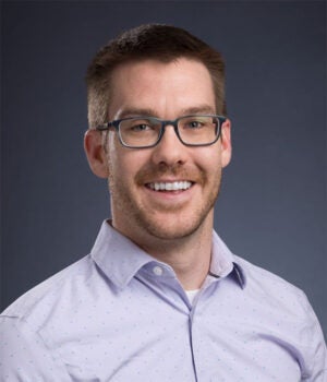 A man with short brown hair, a trimmed beard, and glasses is smiling and wearing a light purple button-up shirt against a plain dark background.