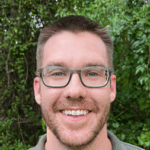 A man with short brown hair, glasses, and a trimmed beard smiles outdoors in front of leafy green trees.