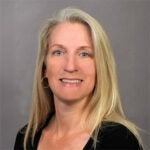 A woman with long blonde hair, wearing a black top and hoop earrings, smiles at the camera against a plain gray background.