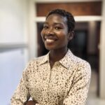 A person with short hair wearing a light-colored, leopard-print button-up shirt stands indoors, smiling with arms crossed.