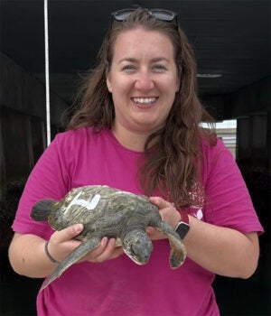 Woman in a pink shirt holds a small sea turtle with a white marking on its shell, smiling at the camera inside an indoor facility.