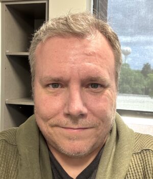 A man with short, light hair and facial stubble is indoors, wearing a green sweater. Behind him are shelves and a window showing trees and a partly cloudy sky.
