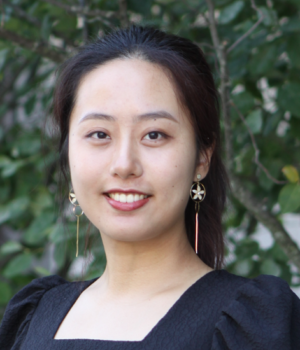 A woman with long dark hair, wearing gold earrings and a black top, is smiling outdoors with leafy green trees in the background.