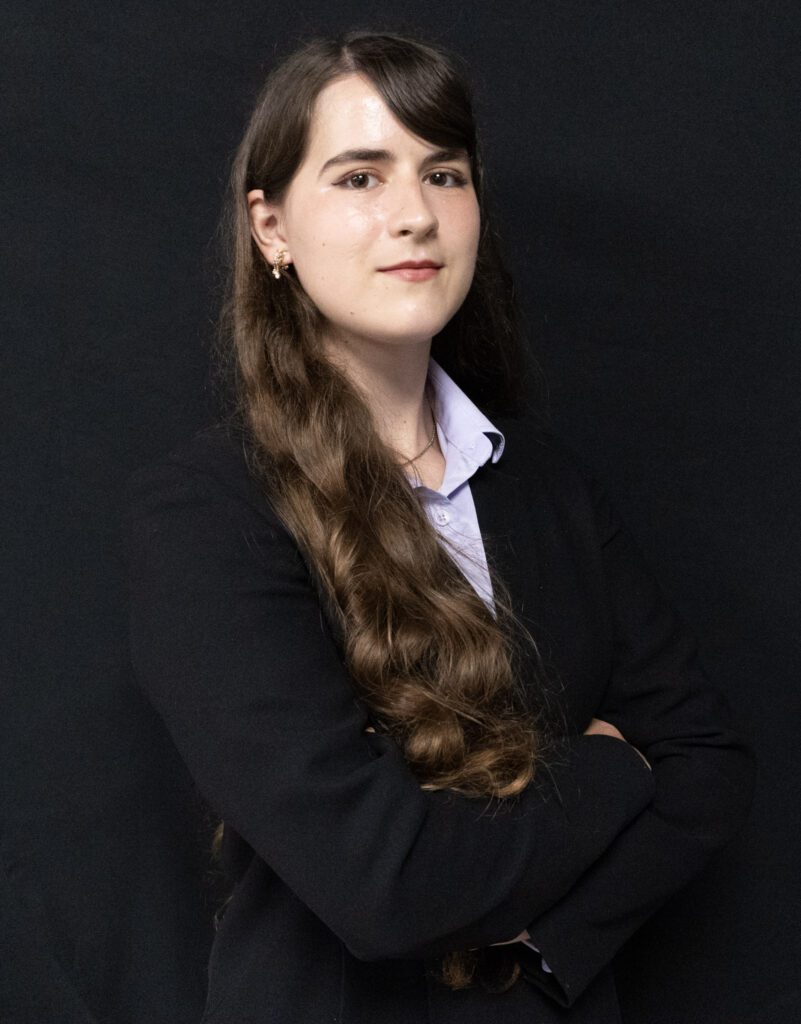 A person with long brown hair wearing a black blazer and light shirt stands with arms crossed in front of a plain black background.