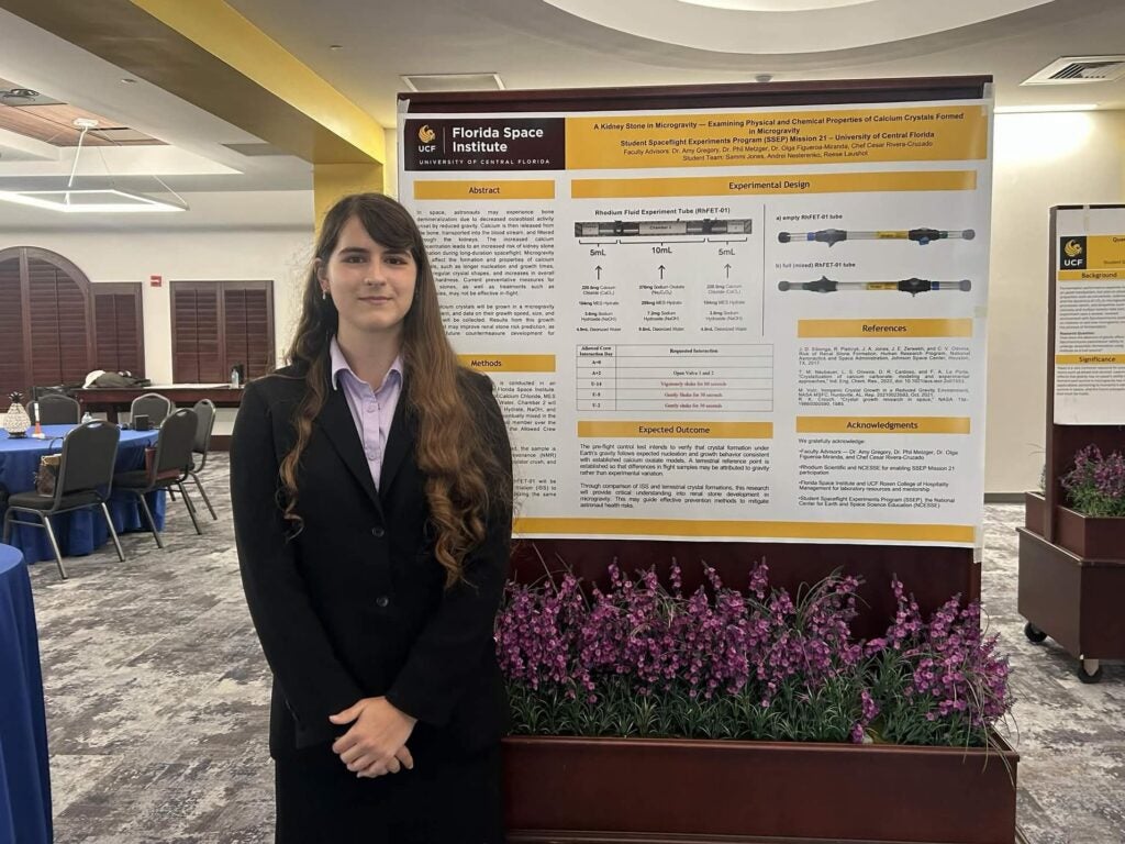 A person in a black suit stands in front of a scientific research poster at a conference room with tables, chairs, and purple flowers.