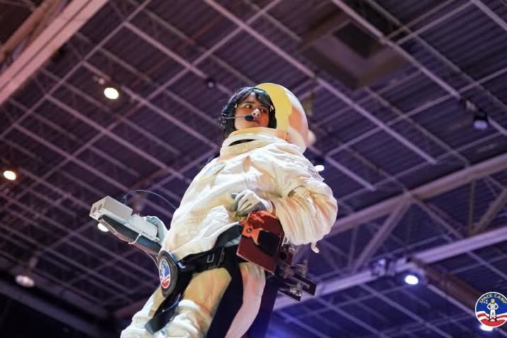 Person in a white spacesuit with a yellow helmet stands indoors under a high metal ceiling, holding equipment, with a NASA logo visible in the corner.