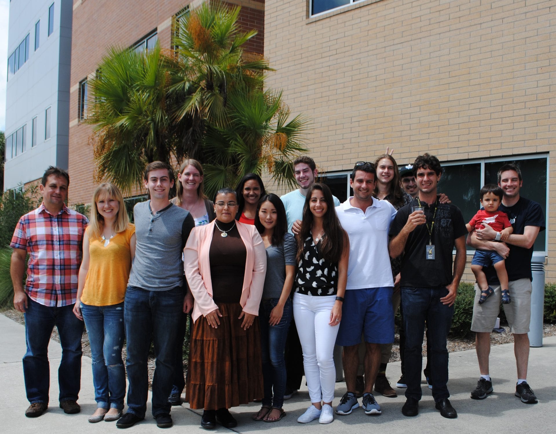 A group of twelve adults and one child standing outdoors in front of a building with palm trees. They are smiling and casually dressed, posing for a group photo.
