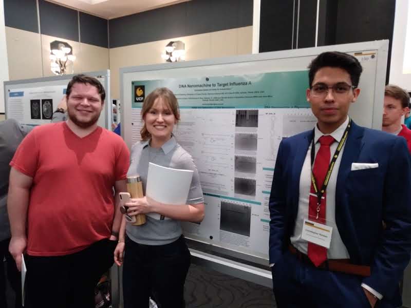 Three individuals stand in front of a scientific poster presentation on DNA nanomachine for Target Influenza A. One person wears a red shirt, another holds a folder, and the third is dressed in a suit.