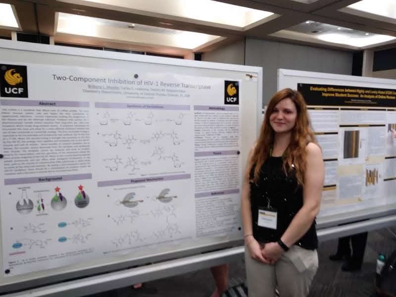 A woman stands in front of a scientific poster titled "Two-Component Inhibition of HIV-1 Reverse Transcriptase" in a conference setting. She is wearing a black top, beige pants, and a name badge.