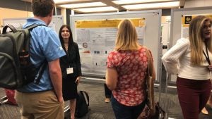 A group of people are gathered around a research poster presentation at a conference. One person stands near the poster, explaining it to others. Two display boards are visible in the background.