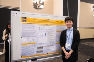 A person stands beside a large presentation poster about sci-fi high alt. The poster is mounted on a freestanding board. Another person is in the background, partially visible.