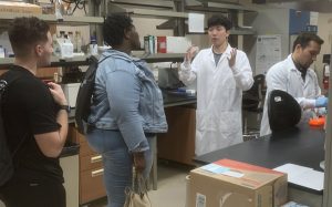 A group of people wearing lab coats and casual clothes are in a lab. One person in a lab coat is speaking while others listen attentively. Lab equipment and supplies are visible in the background.
