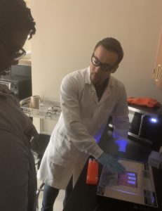 A person wearing a lab coat and gloves explains a procedure to another individual in a laboratory setting.