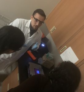 A person in a lab coat and safety glasses demonstrates a procedure to two individuals in a laboratory setting.