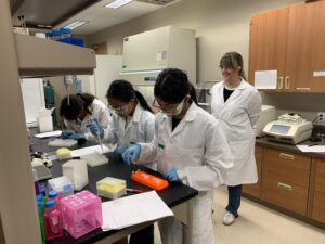 Four people in lab coats and safety glasses work in a laboratory; three are using pipettes at a bench while one stands observing in the background.