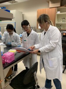 Three people in lab coats work together at a laboratory bench, one holding a clipboard and discussing notes while the others handle equipment and samples.