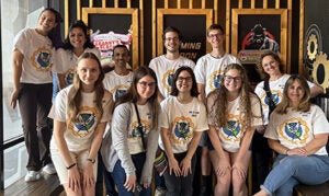 A group of eleven people wearing matching white graphic t-shirts pose together indoors, smiling for the camera in front of a decorated wall.