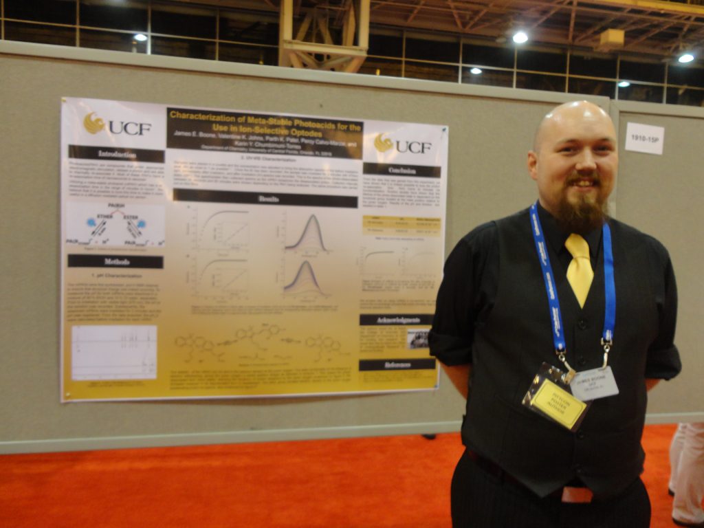 A man in a black suit and yellow tie presenting a scientific poster at a conference.
