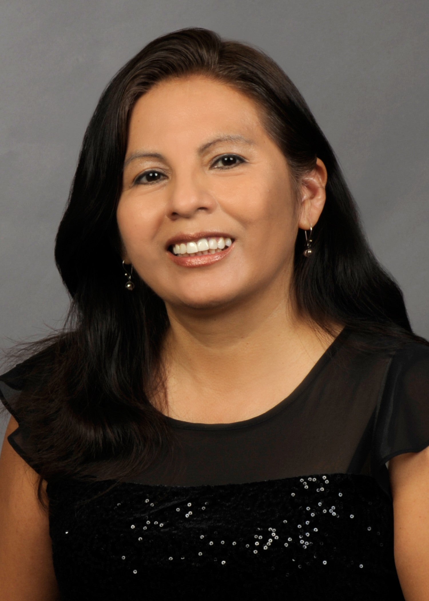 thumbnail_Karin YCHT Picture Portrait of a smiling woman with long dark hair, wearing a black sequined top and earrings.