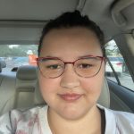 Skylar 2024 A person with glasses and a floral top sits inside a car, smiling slightly. The parking lot can be seen through the windows.