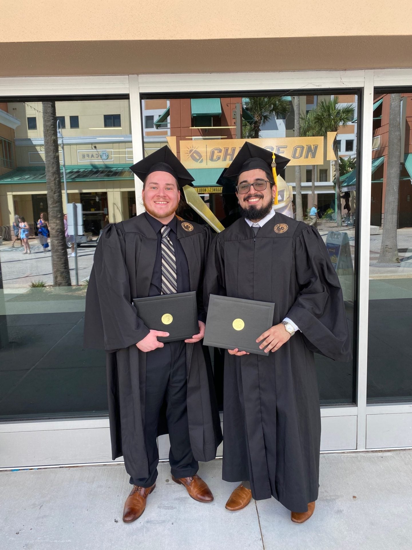 Two graduates in black caps and gowns stand side by side holding diplomas in front of a glass building with a sign that reads "Charge On.