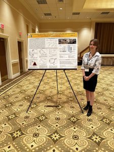 A person stands beside an academic poster displayed on an easel in a conference room. The room features patterned carpet, beige walls, and double doors.