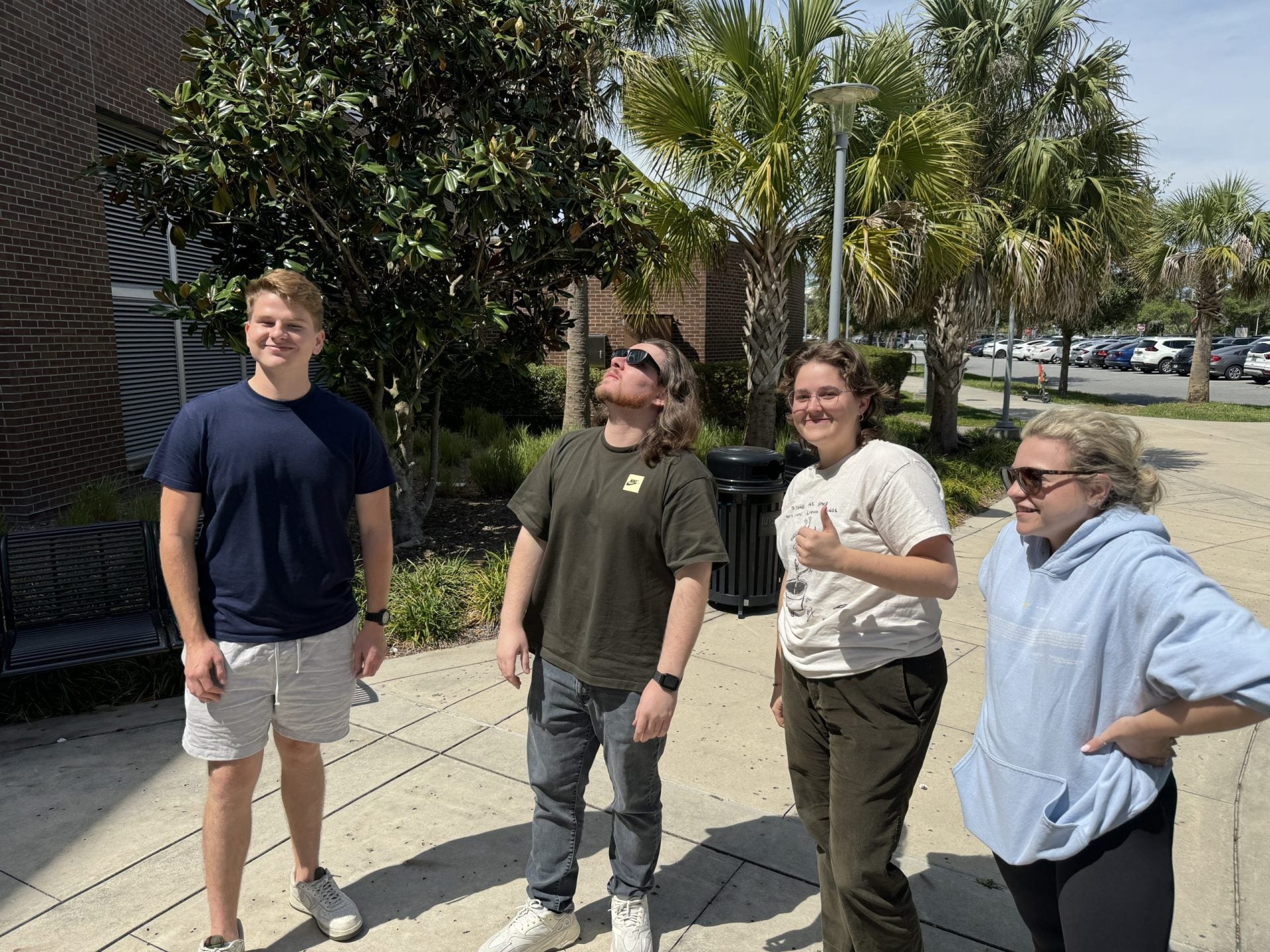 Four people standing on a sidewalk outside on a sunny day, with trees and a brick building in the background. One person is looking up with sunglasses, and another is giving a thumbs-up gesture.