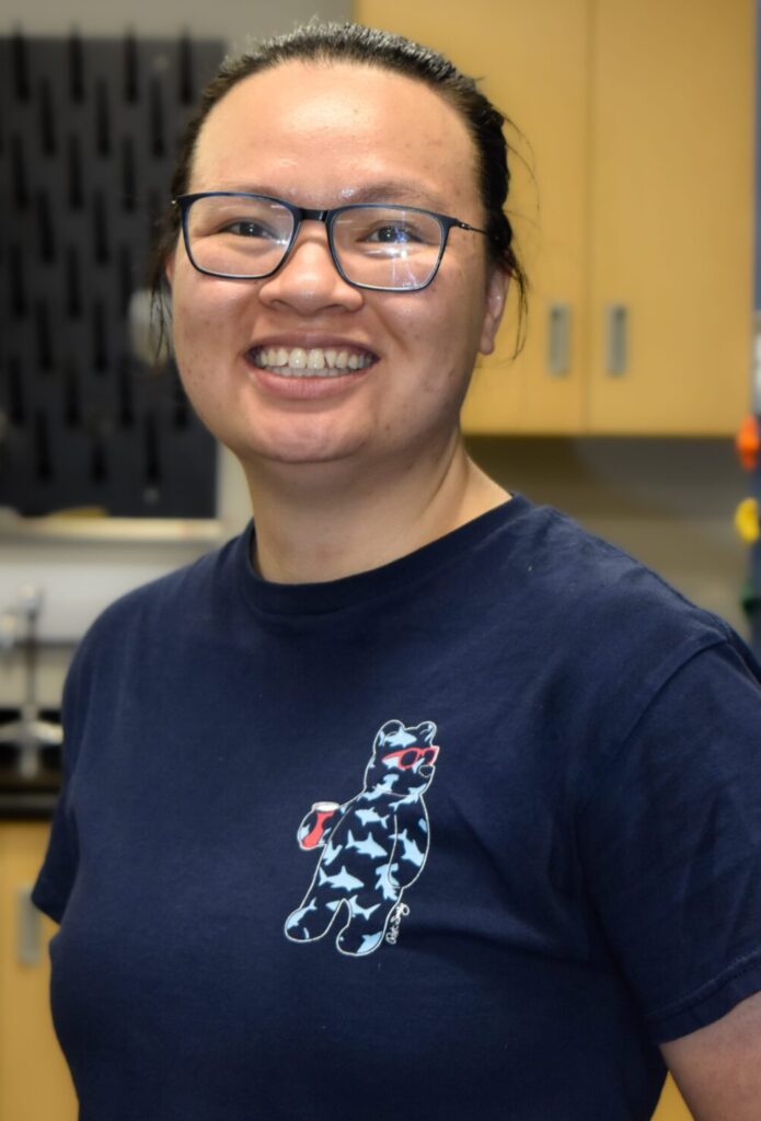 Person wearing glasses and a navy blue T-shirt with a cat graphic, smiling in an indoor setting with cabinets and equipment in the background.