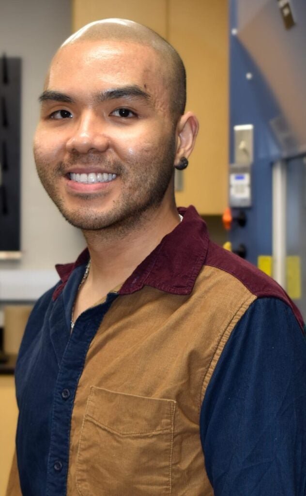 Person smiling in a laboratory setting, wearing a multicolored collared shirt. Laboratory equipment and cabinets are visible in the background.