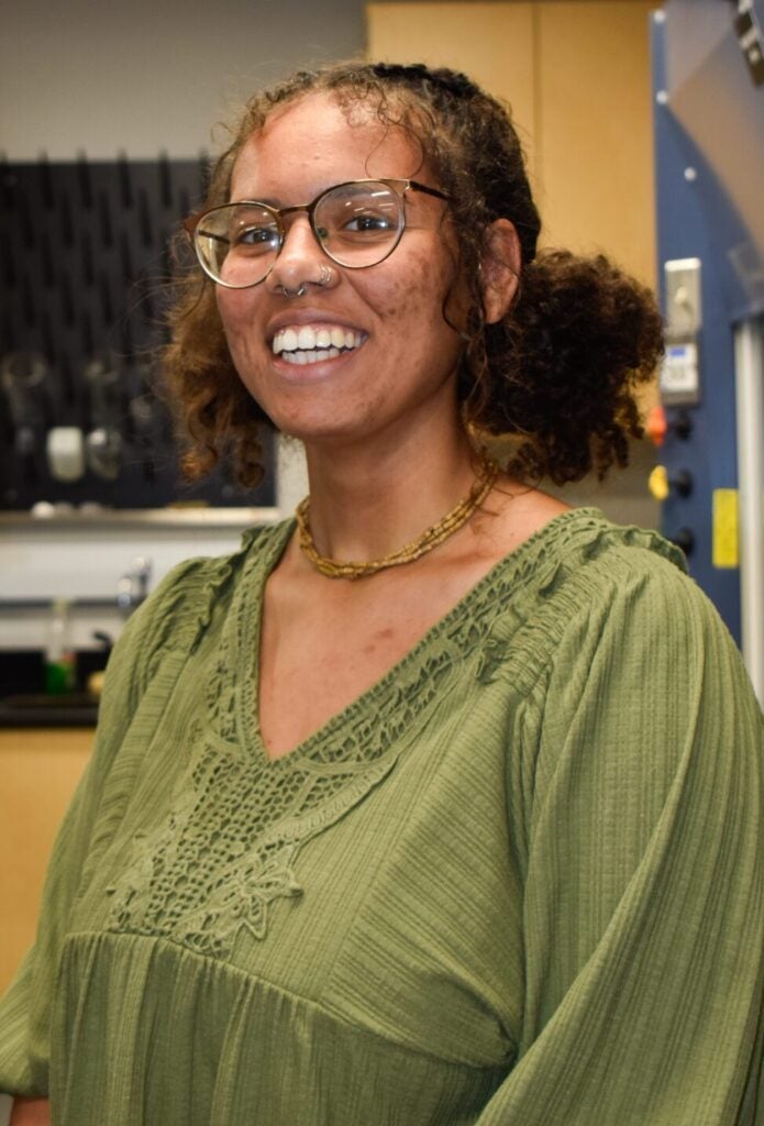 A person wearing glasses and a green blouse stands smiling in a laboratory setting with equipment and cabinets in the background.