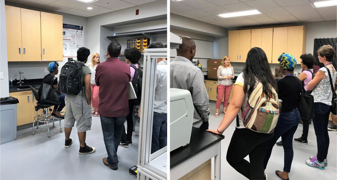 A group of people stands in a lab, listening to a woman speak; lab equipment and cabinets are visible in the background.
