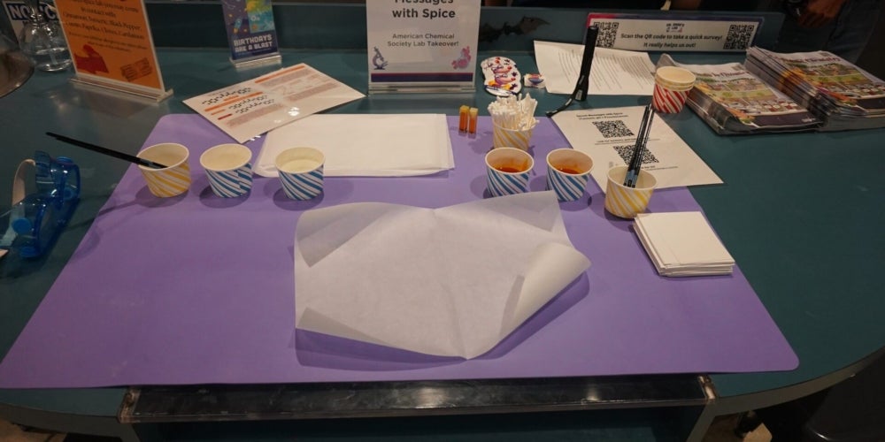 Table setup for a “Secret Messages with Spice” activity, with cups, brushes, paper, and informational materials arranged on a purple mat.