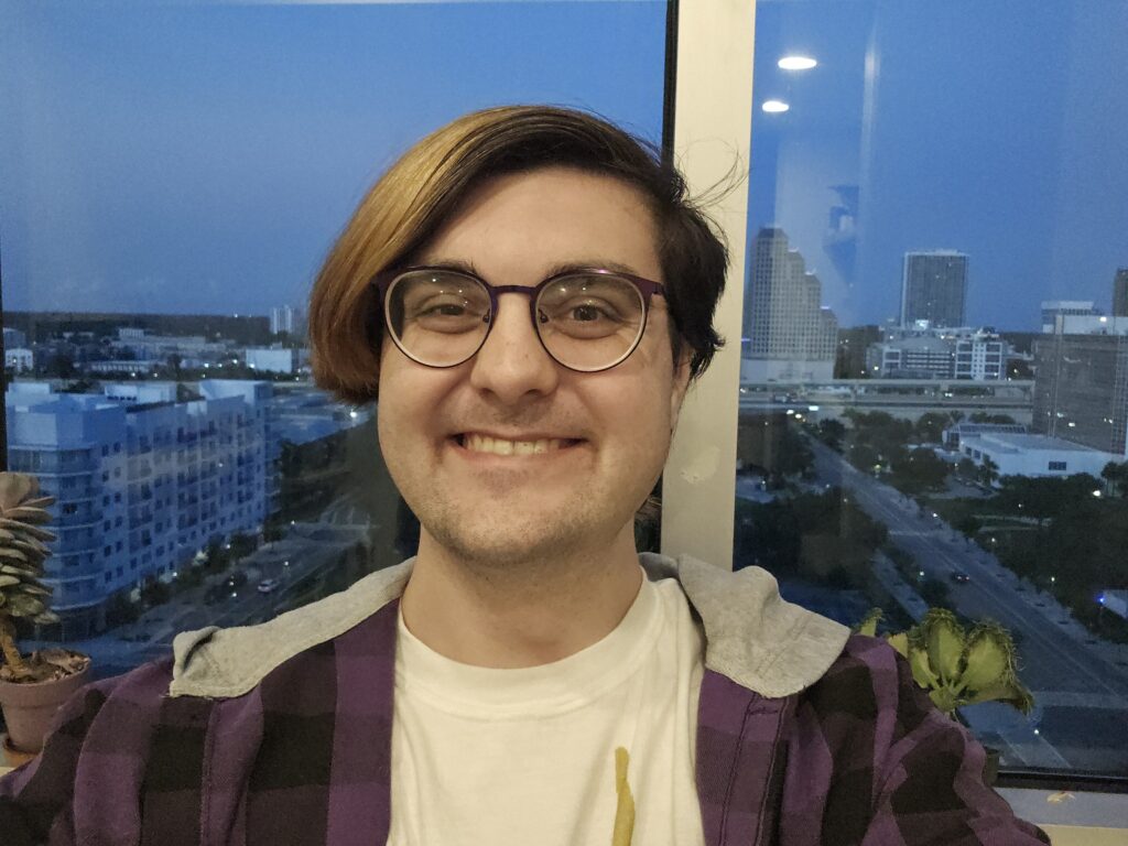 A person with glasses and a purple plaid shirt smiles at the camera indoors with a cityscape visible through a window behind them.