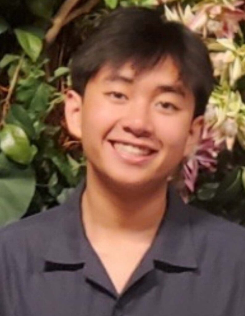 A young person with short dark hair smiles at the camera, wearing a dark collared shirt, with green leaves and flowers in the background.