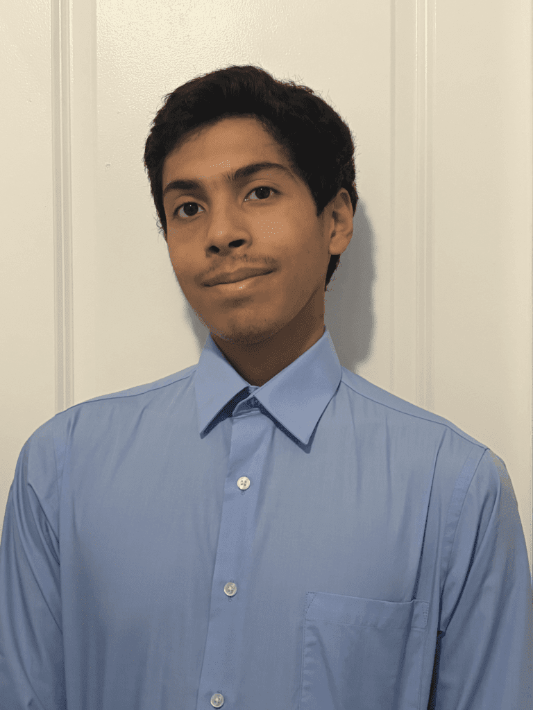 A young person with short dark hair wearing a light blue button-up shirt stands in front of a white wall, looking at the camera.
