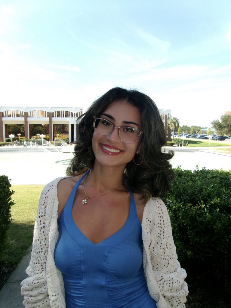 Woman with shoulder-length dark hair, wearing glasses, a blue dress, and a white cardigan, stands smiling outdoors with buildings, bushes, and a fountain in the background under a blue sky.